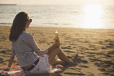 Seitenansicht einer Frau, die eine Bierflasche hält, während sie sich am Strand bei Sonnenuntergang entspannt - CAVF70656