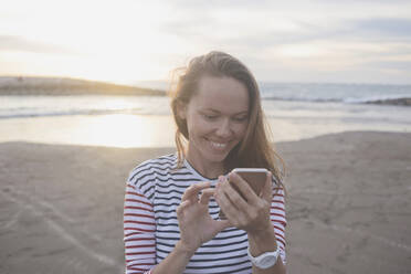Smiling woman using mobile phone at beach during sunset - CAVF70660