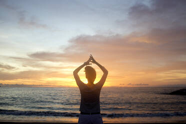 Rückansicht einer Frau mit erhobenen Armen, die bei Sonnenuntergang am Strand steht - CAVF70664