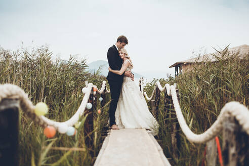 Couple embracing while standing on footbridge amidst plants - CAVF70736
