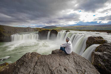 Man sitting on edge of rock, Godafoss waterfall, BᲰardalur, Iceland - CUF53969