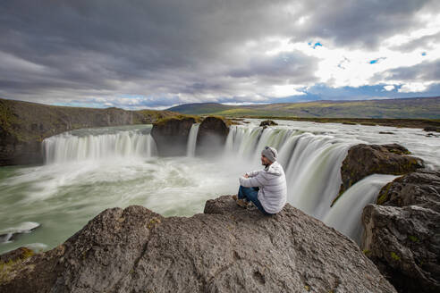 Mann sitzt am Rand eines Felsens, Godafoss-Wasserfall, BᲰardalur, Island - CUF53969