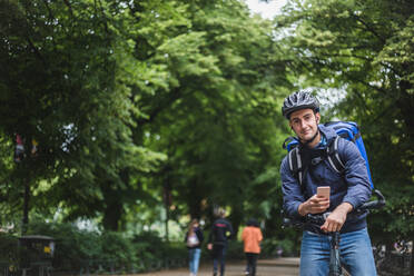 Porträt eines selbstbewussten Zustellers mit Fahrrad vor einem Baum in der Stadt - MASF15279