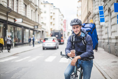 Porträt eines selbstbewussten Lebensmittellieferanten mit Fahrrad auf einer Straße in der Stadt, lizenzfreies Stockfoto