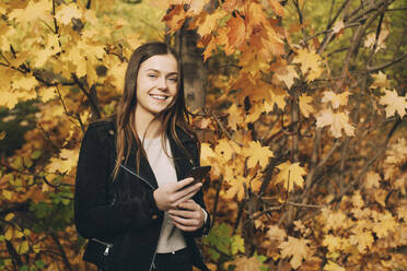 Portrait of smiling teenage girl with mobile phone standing against trees during autumn - MASF15360
