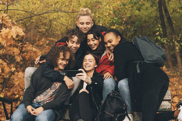 Teenage girl taking selfie with friends while sitting on bench against trees during autumn - MASF15367