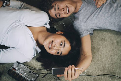 Glücklicher Teenager, der ein Selfie mit einem Freund auf dem Handy macht, während er auf dem Boden im Wohnzimmer liegt - MASF15396