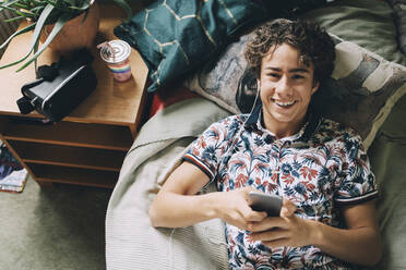 Hochwinkliges Porträt eines glücklichen Teenagers, der auf dem Bett zu Hause Musik über sein Smartphone hört - MASF15412