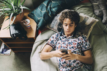 Hoher Blickwinkel eines männlichen Teenagers, der mit seinem Handy Musik hört, während er zu Hause auf dem Bett liegt - MASF15415
