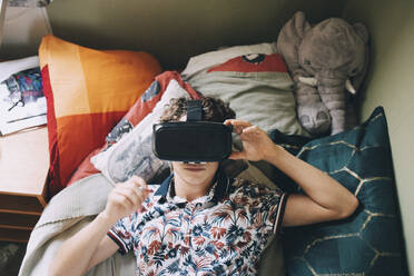 Teenage boy wearing virtual reality headset while lying on bed at home - MASF15417