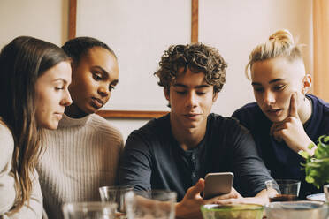 Teenager, der Freunden sein Handy zeigt, während er am Tisch im Zimmer sitzt - MASF15445