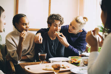 Teenager füttert einen Freund mit Pizza, während die Frauen sie im Wohnzimmer betrachten - MASF15450