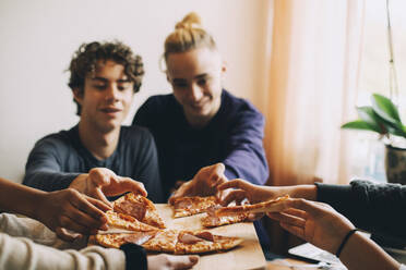 Male and female teenage friends taking slices of pizza from tray at home - MASF15452