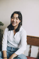 Portrait of smiling teenage girl with braces sitting on sofa against wall at home - MASF15456