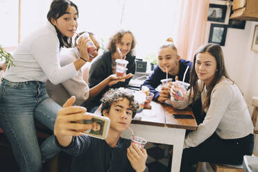 Teenage boy taking selfie with friends through mobile phone while enjoying smoothie at home - MASF15468