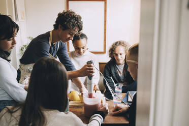 Freunde schauen auf einen Teenager, der am Esstisch im Zimmer einen Smoothie zubereitet - MASF15469