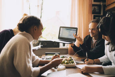 Smiling teenage boy showing laptop to friends while studying at table - MASF15497