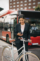 Lächelnde Geschäftsfrau, die ihr Smartphone benutzt, während sie mit dem Fahrrad gegen einen Bus auf der Straße in der Stadt steht - MASF15506