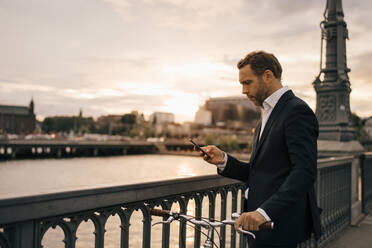 Businessman using mobile phone while standing with bicycle on bridge in city during sunset - MASF15508