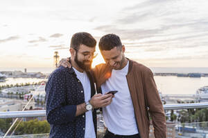 Schwules Paar mit Handy auf einem Aussichtspunkt über der Stadt mit Blick auf den Hafen, Barcelona, Spanien - AFVF04438