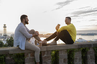 Zwei Männer sitzen auf einer Mauer auf einem Aussichtspunkt über der Stadt mit Blick auf den Hafen, Barcelona, Spanien - AFVF04447