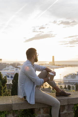 Mann sitzt auf einer Mauer auf einem Aussichtspunkt über der Stadt mit Blick auf den Hafen, Barcelona, Spanien, lizenzfreies Stockfoto