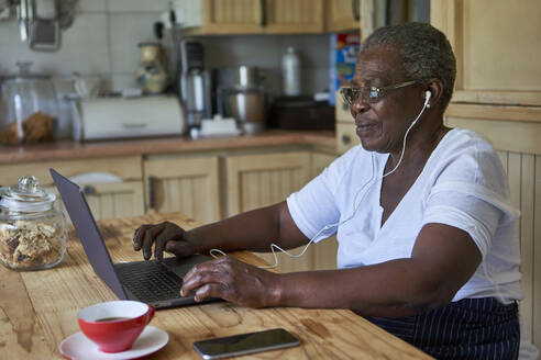 Ältere Frau sitzt am Küchentisch und benutzt einen Laptop - VEGF01079