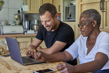 Senior woman and smiling man sitting at kitchen table sharing laptop - VEGF01088