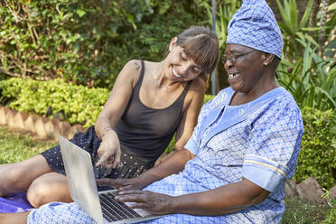 Happy senior woman sitting on lawn sharing laptop with a woman - VEGF01090