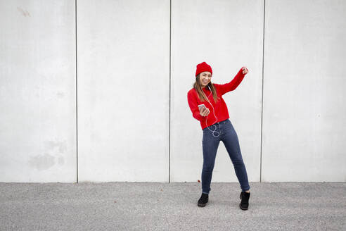 Woman wearing red pullover and wolly hat and dancing in front of a wall - HMEF00691