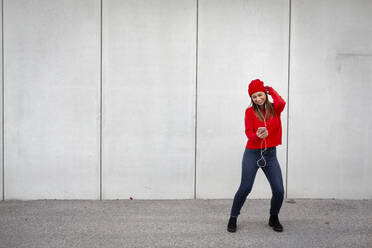 Woman wearing red pullover and wolly hat and dancing in front of a wall - HMEF00692