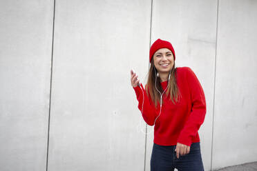 Woman wearing red pullover and wolly hat and dancing in front of a wall - HMEF00693