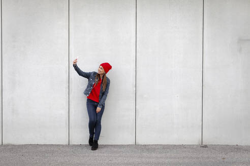 Woman wearing red pullover and wolly hat and taking a selfie in front of a wall - HMEF00699