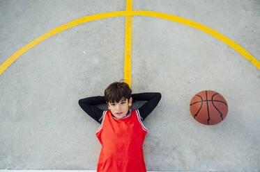 Porträt eines Jungen, der mit einem Basketball auf einem Platz im Freien liegt - JCMF00341