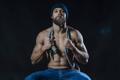 Porträt eines barbusigen Sportlers mit Springseil im Studio sitzend - DLTSF00316