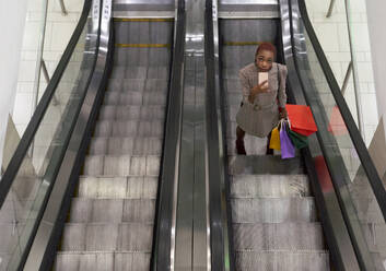 Junge Frau mit bunten Einkaufstüten in der Hand und mit ihrem Telefon auf der Rolltreppe stehend - VEGF01135