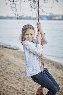 Porträt eines lächelnden kleinen Mädchens, das am Flussufer mit einem Seil schwingt - RORF01970