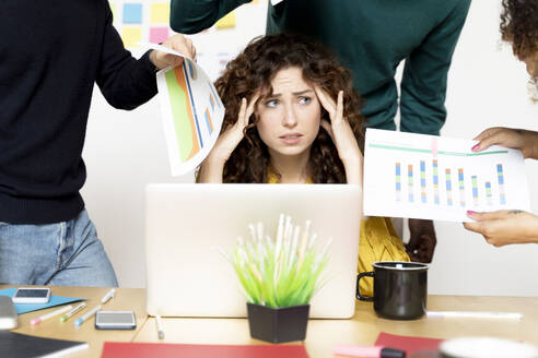 Stressed woman siting at desk in office surrounded by colleagues - FMOF00831