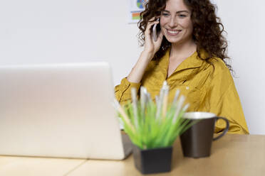 Smiling woman using laptop and smartphone at desk in office - FMOF00836