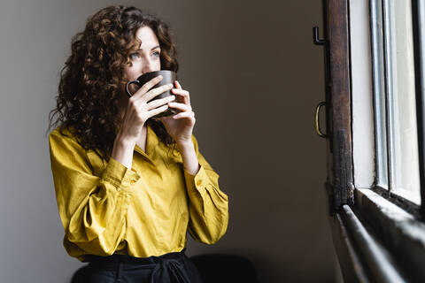 Frau schaut aus dem Fenster und trinkt aus einer Kaffeetasse, lizenzfreies Stockfoto