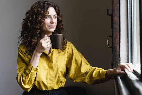 Frau schaut mit Kaffeetasse aus dem Fenster, lizenzfreies Stockfoto