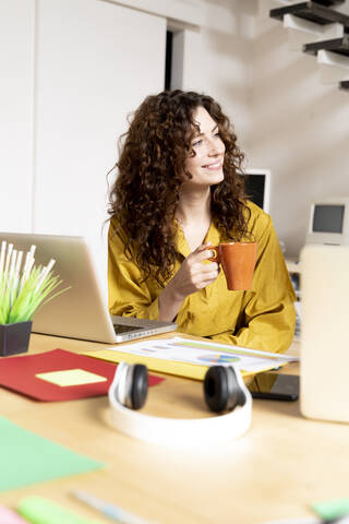 Lächelnde Frau mit einer Tasse Kaffee am Schreibtisch sitzend im Büro, lizenzfreies Stockfoto