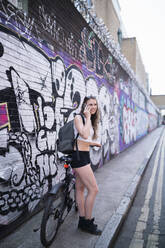 Lächelnde junge Frau am Telefon an einer Graffitiwand - FBAF01090