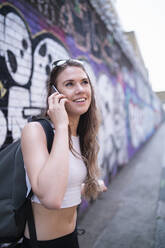 Lächelnde junge Frau am Telefon an einer Graffitiwand - FBAF01091