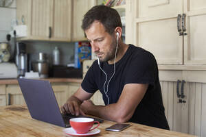 Mann arbeitet von zu Hause aus, sitzt am Küchentisch, benutzt Laptop und Smartphone - VEGF01212
