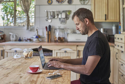 Man working from home, sitting at kitchen table, using laptop and smartphone stock photo