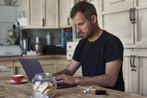 Mann arbeitet von zu Hause aus, sitzt am Küchentisch, benutzt Laptop und Smartphone - VEGF01219