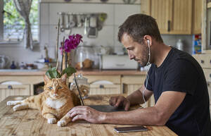 Mann arbeitet von zu Hause aus, sitzt mit Katze am Küchentisch und benutzt einen Laptop - VEGF01221