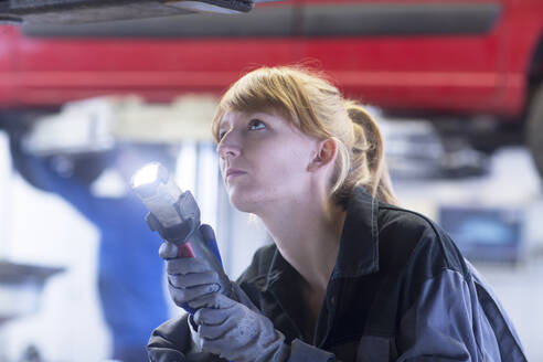 Weiblicher Automechaniker mit Lampe, der auf den Boden eines Autos in einer Reparaturwerkstatt schaut - SGF02531