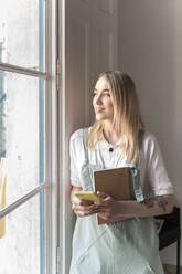 Junge Frau mit Notizbuch und Handy schaut aus dem Fenster - AFVF04465